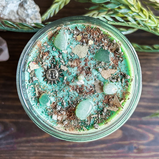Green intention crystal candle that is decorated with a witchy sun and moon charm, gold magic glitter. The candle also has crystals and herbs for manifesting abundance.