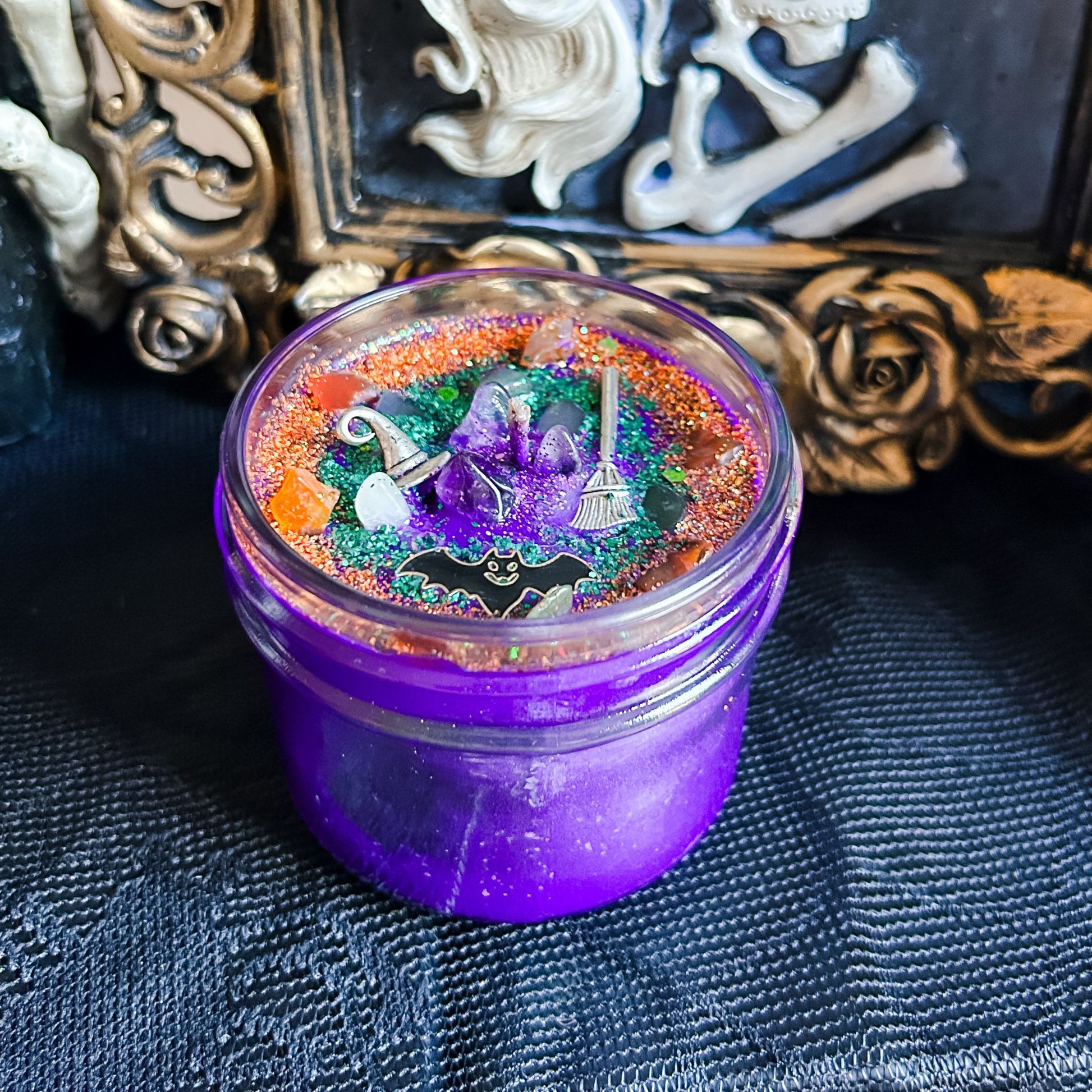 A purple Halloween candle that is decorated with a witch's broom, a witch hat, a spooky bat, witchy crystals, and magic glitter.
