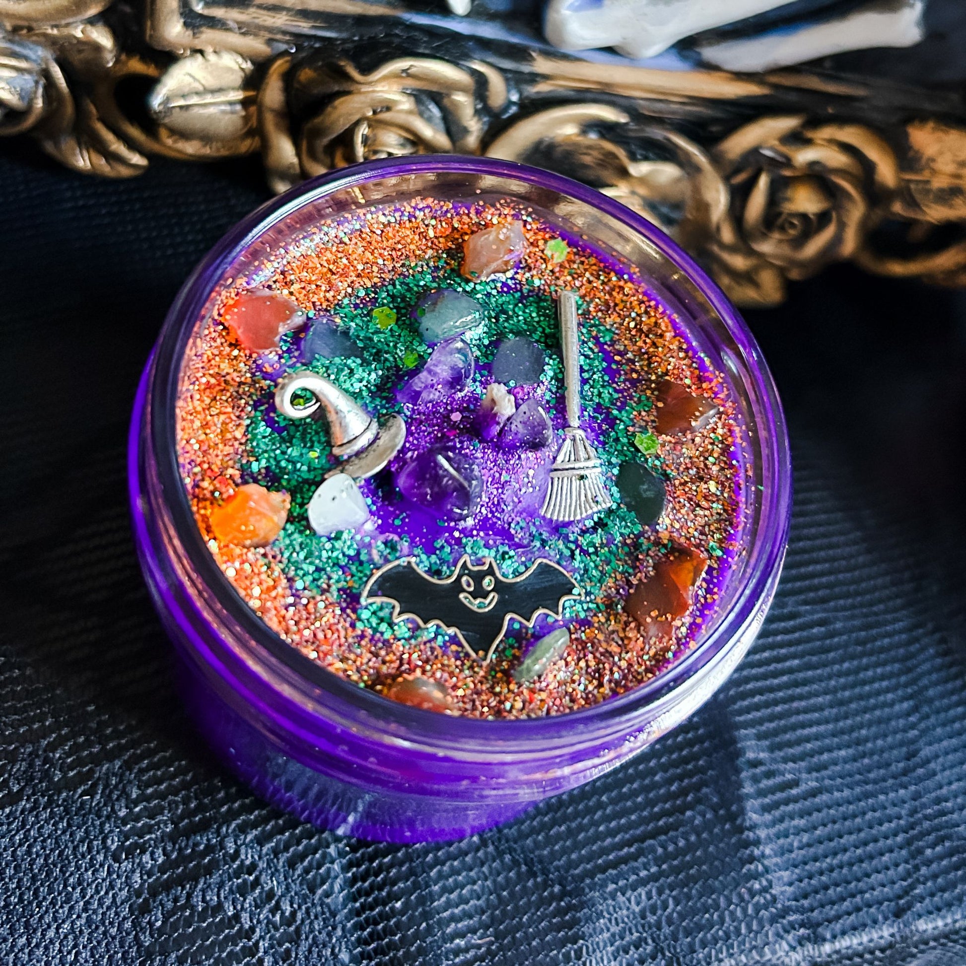 A purple Halloween candle that is decorated with a witch's broom, a witch hat, a spooky bat, witchy crystals, and magic glitter.