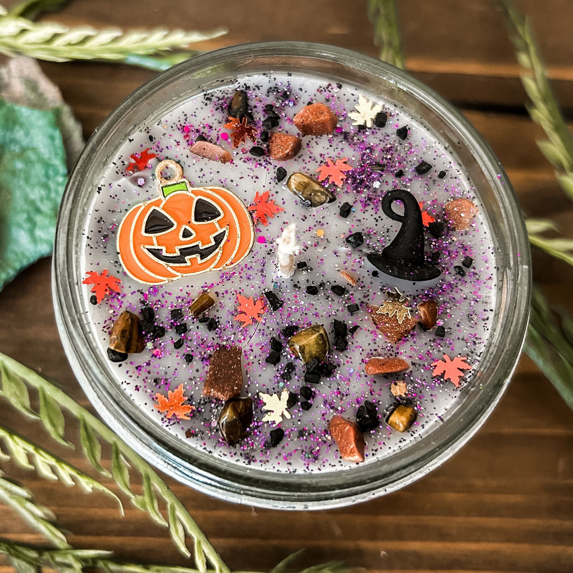 A Halloween candle that is decorated with a jack o lantern, a witch hat, spooky crystals, and autumn glitter.