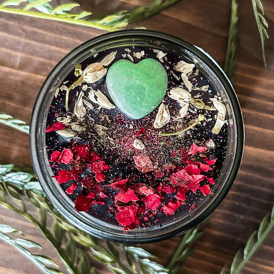Black intention candle that is decorated with a green aventurine crystal heart. The candle also has jasmine, rose petals, witch hazel, and magic glitter for manifesting.