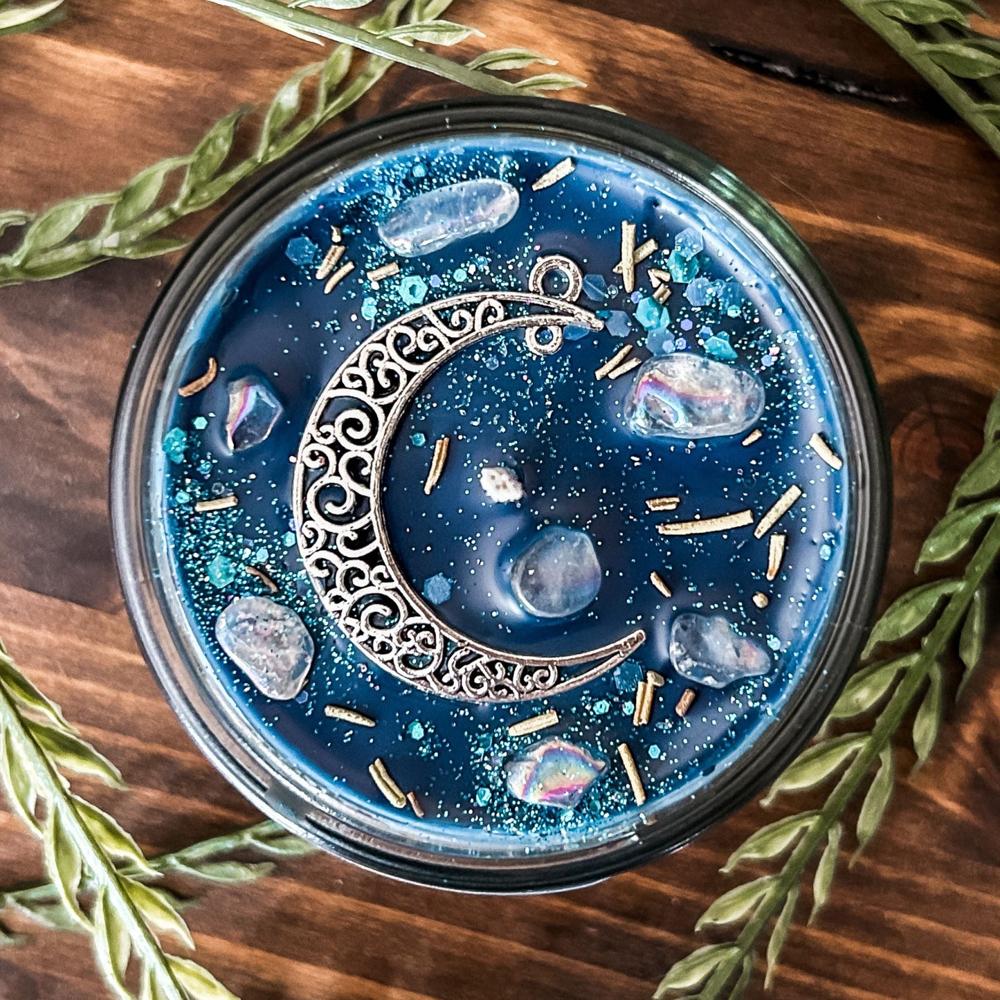 Blue intention candle that is decorated with a silver crescent moon. The candle also has rosemary, aura quartz crystals, and celestial glitter for moon ritual spells and manifesting.