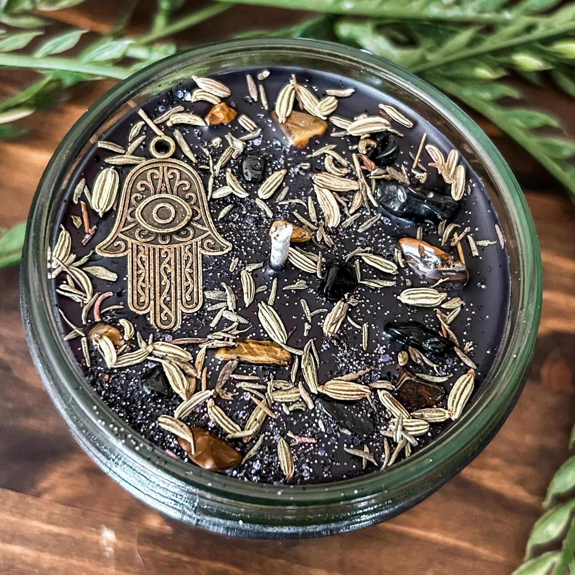 Black crystal intention candle that is decorated with a Hamsa hand, tiger's eye crystal, black tourmaline crystal, thyme, fennel seed, and witchy glitter for protection and grounding.