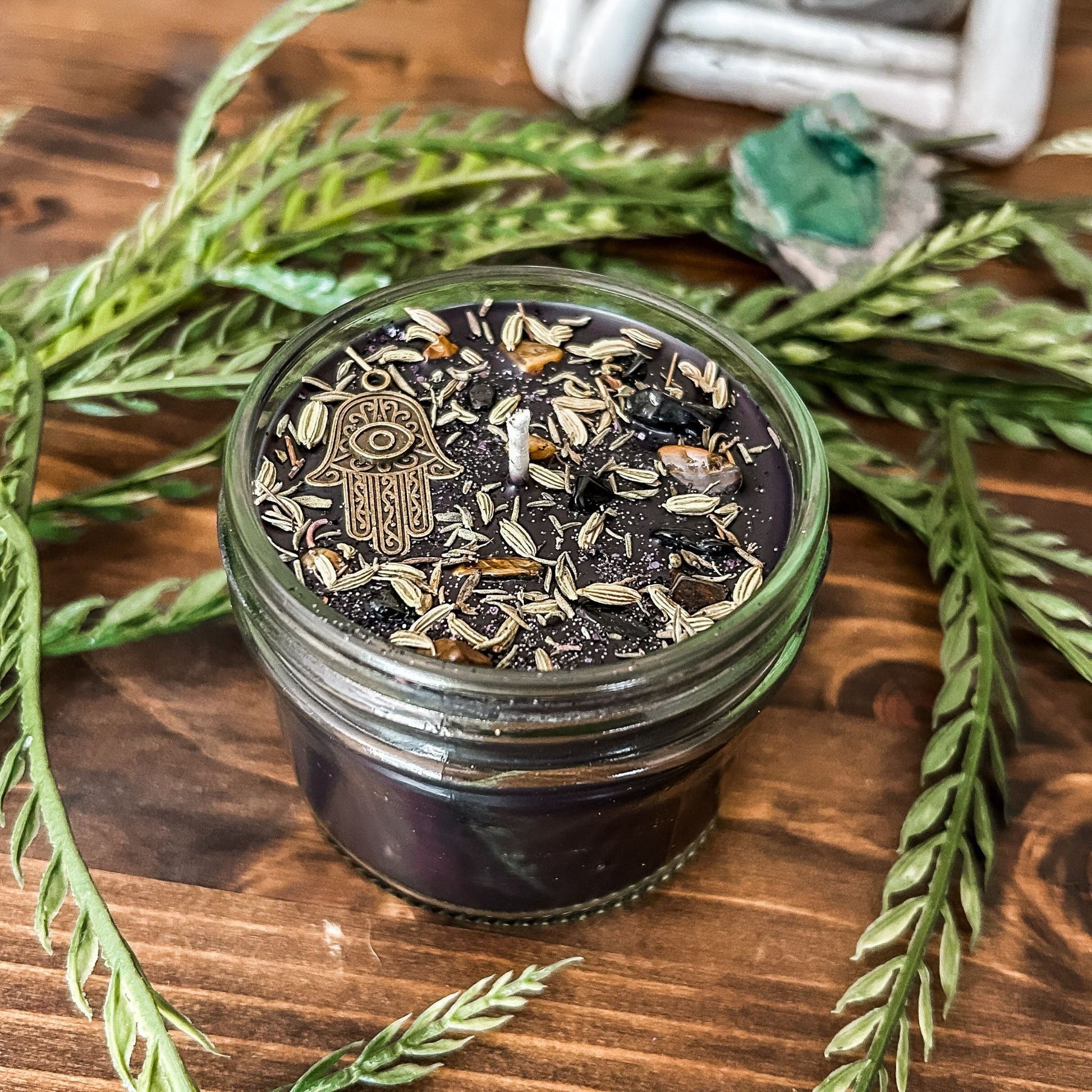 Black crystal intention candle that is decorated with a Hamsa hand, tiger's eye crystal, black tourmaline crystal, thyme, fennel seed, and witchy glitter for protection and grounding.