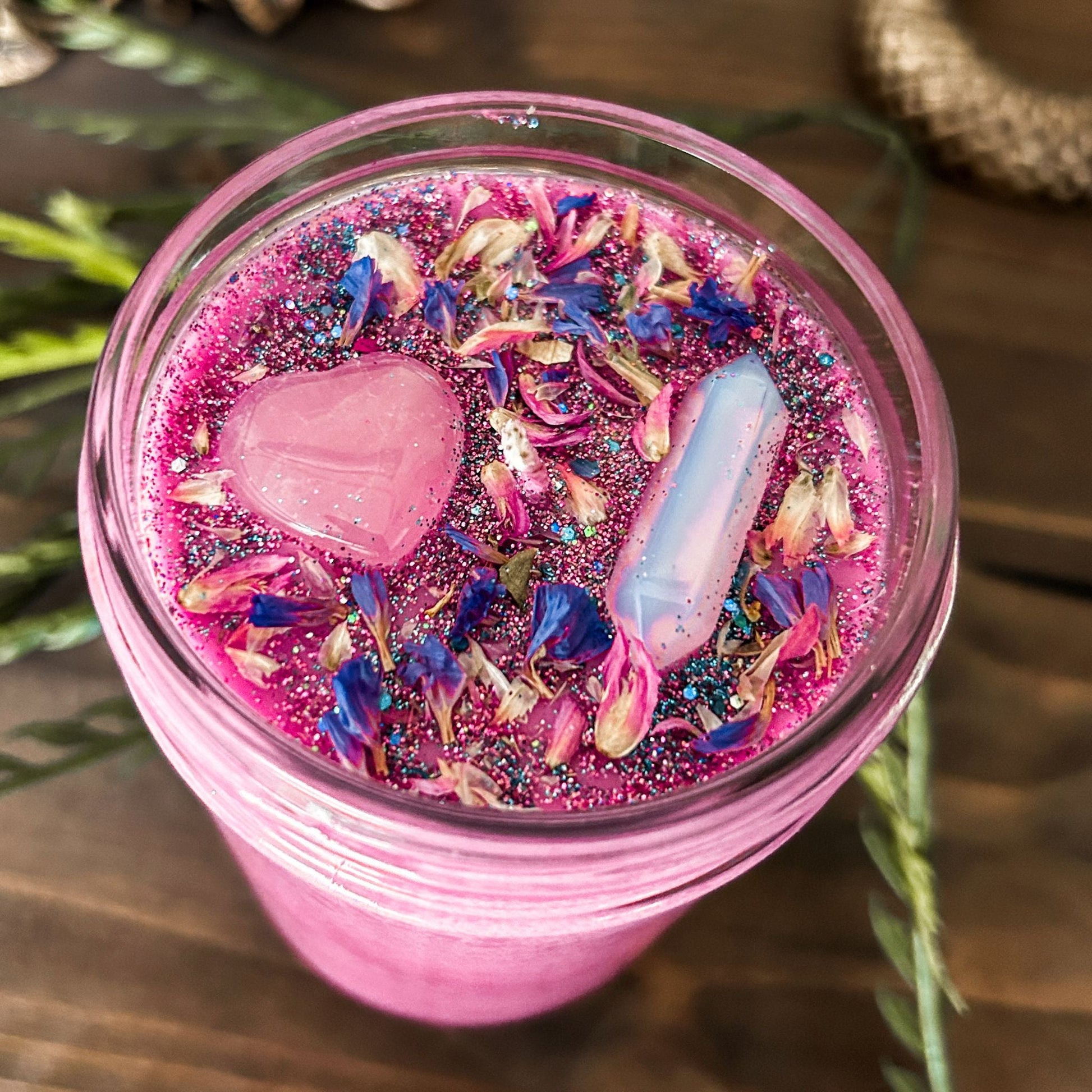 Bright pink witchy intention candle that is decorated with a rose quartz crystal heart, an aura quartz crystal point, flowers, and blue glitter for manifesting self-love and confidence.