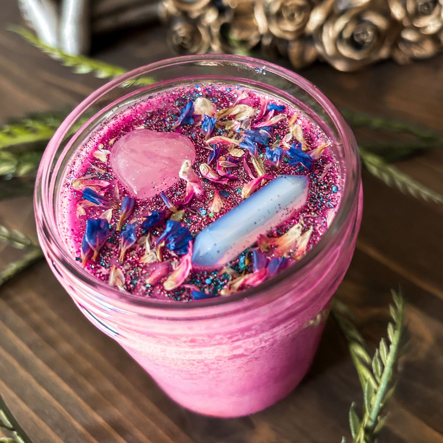 Bright pink witchy intention candle that is decorated with a rose quartz crystal heart, an aura quartz crystal point, flowers, and blue glitter for manifesting self-love and confidence.