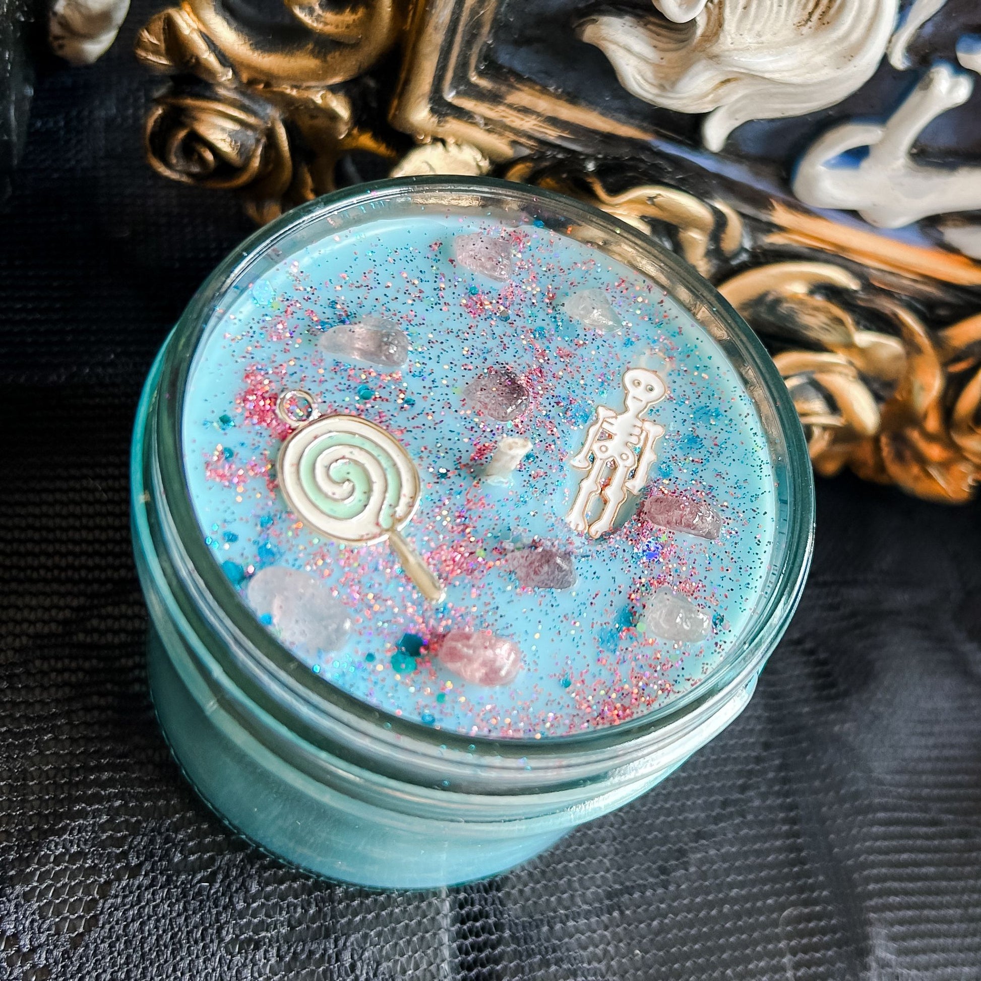 A pastel blue Halloween candle with a creepy cute skeleton, a swirl lollipop, spooky crystals, and gothic pink glitter.