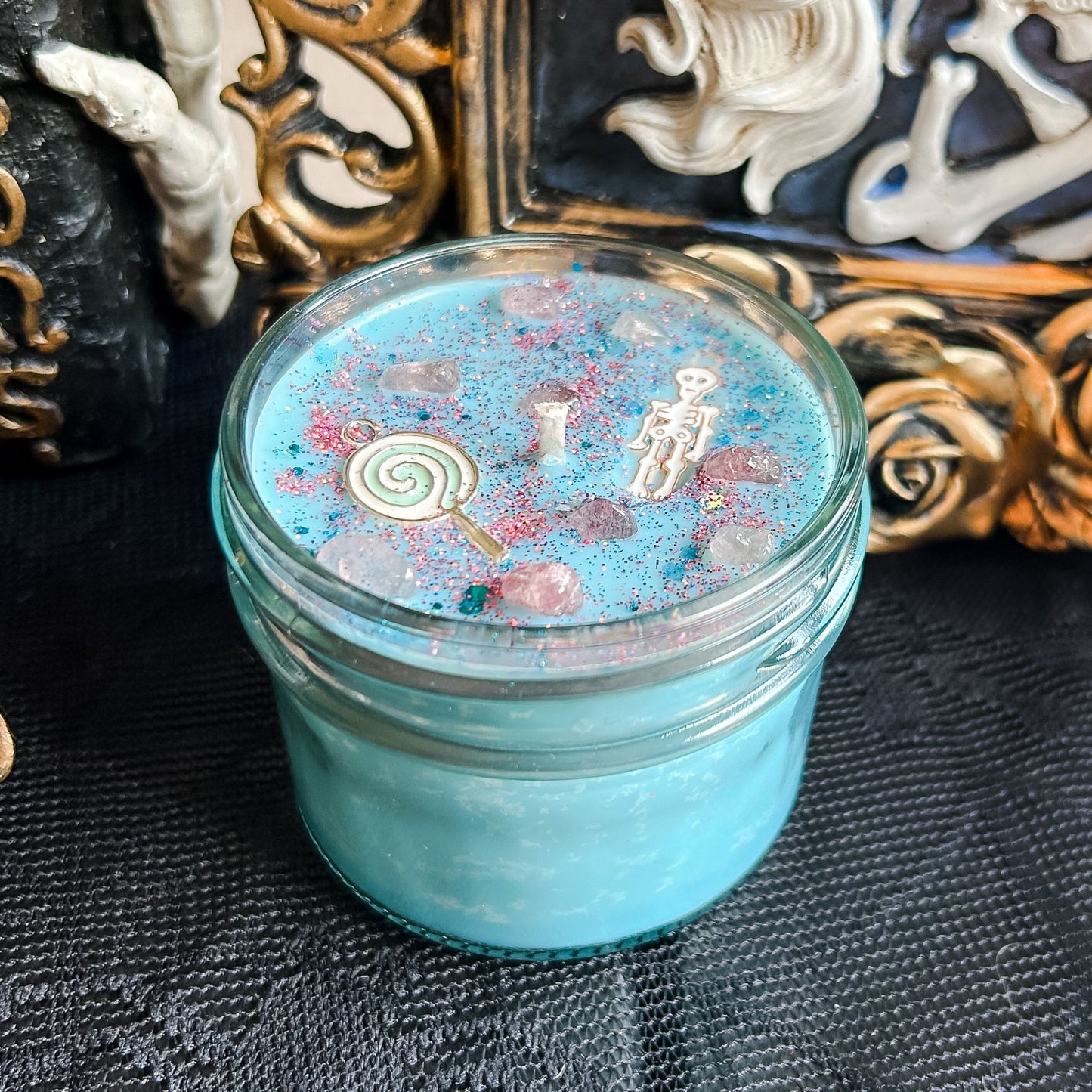 A pastel blue Halloween candle with a creepy cute skeleton, a swirl lollipop, spooky crystals, and gothic pink glitter.