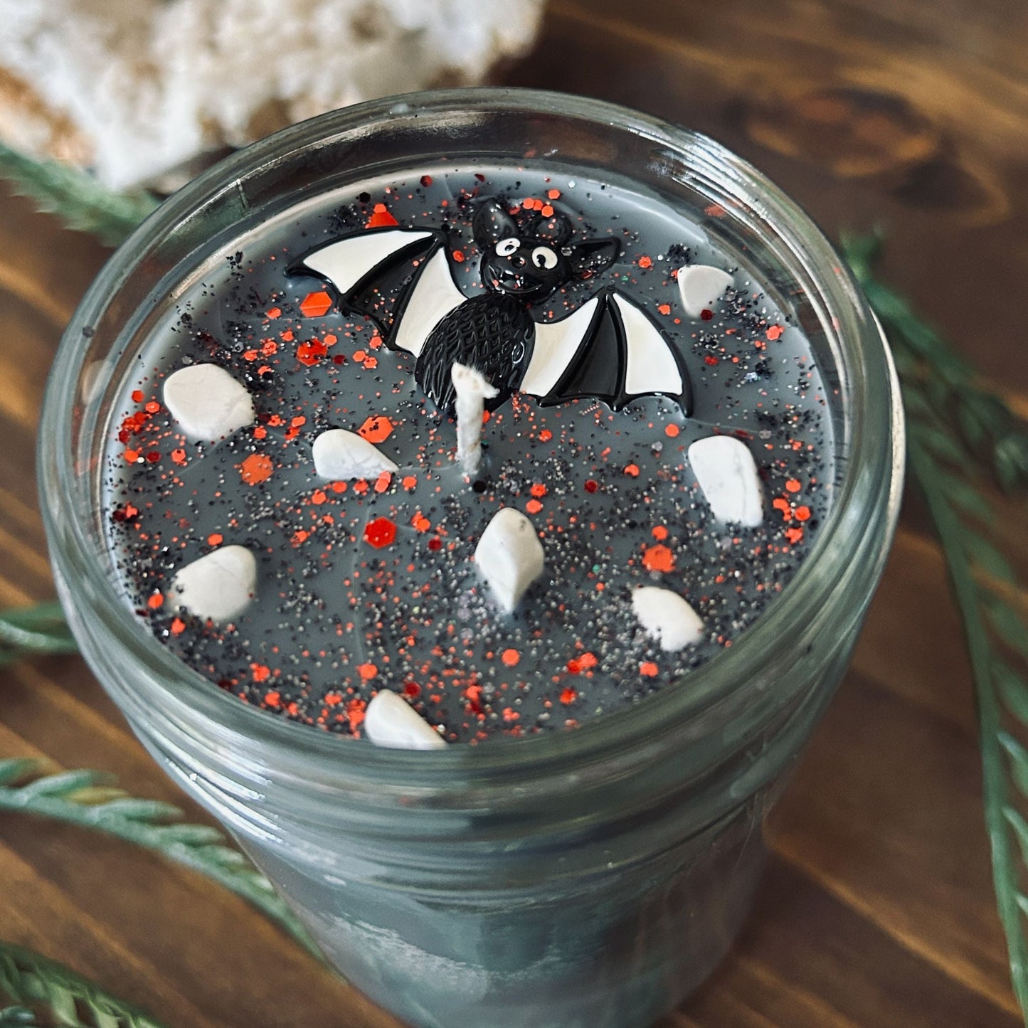 A gray Halloween candle that is decorated with a spooky black and white striped bat charm, white crystals, and gothic glitter.