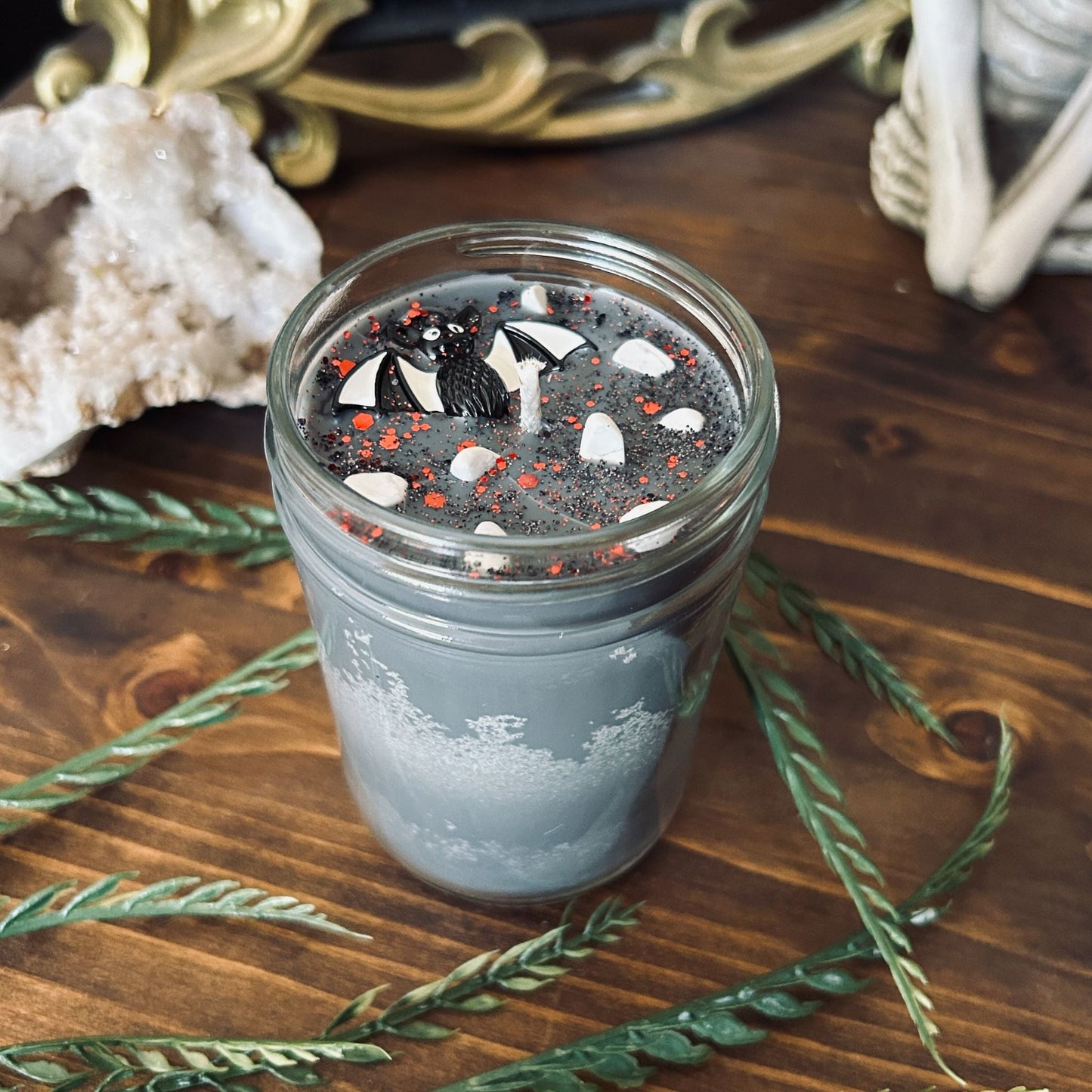 A gray Halloween candle that is decorated with a spooky black and white striped bat charm, white crystals, and gothic glitter.