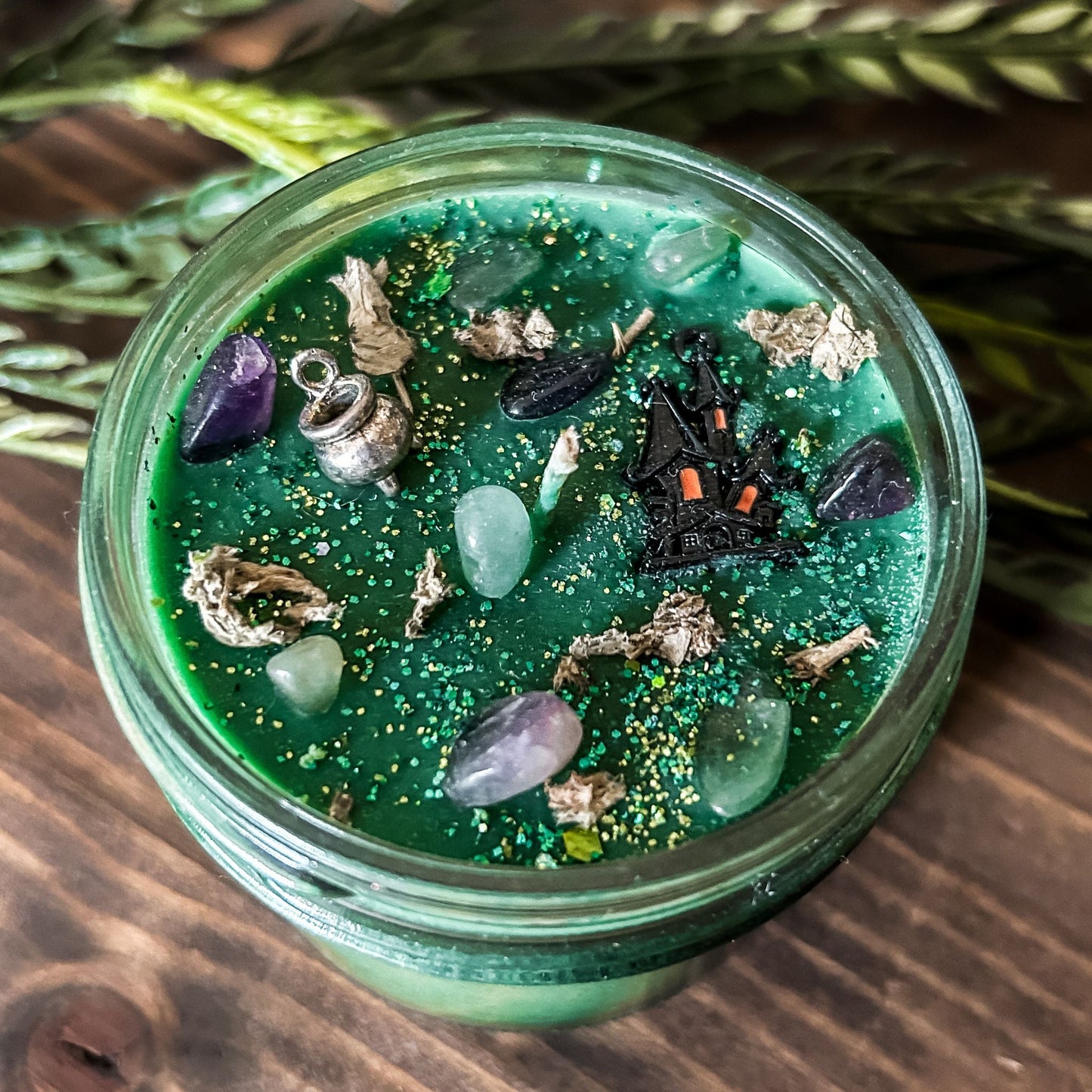 A Halloween candle that is decorated with a haunted house, a cauldron, spooky crystals, and witchy green and purple glitter.