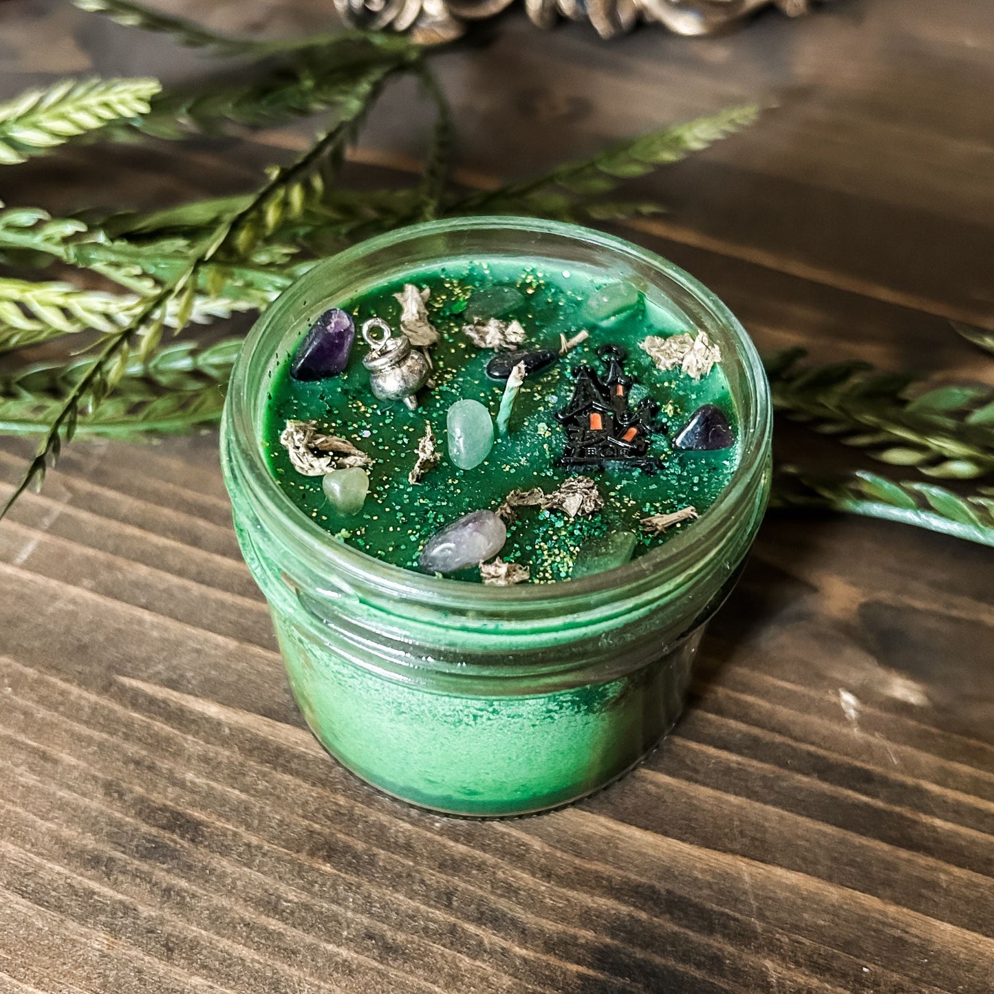 A Halloween candle that is decorated with a haunted house, a cauldron, spooky crystals, and witchy green and purple glitter.