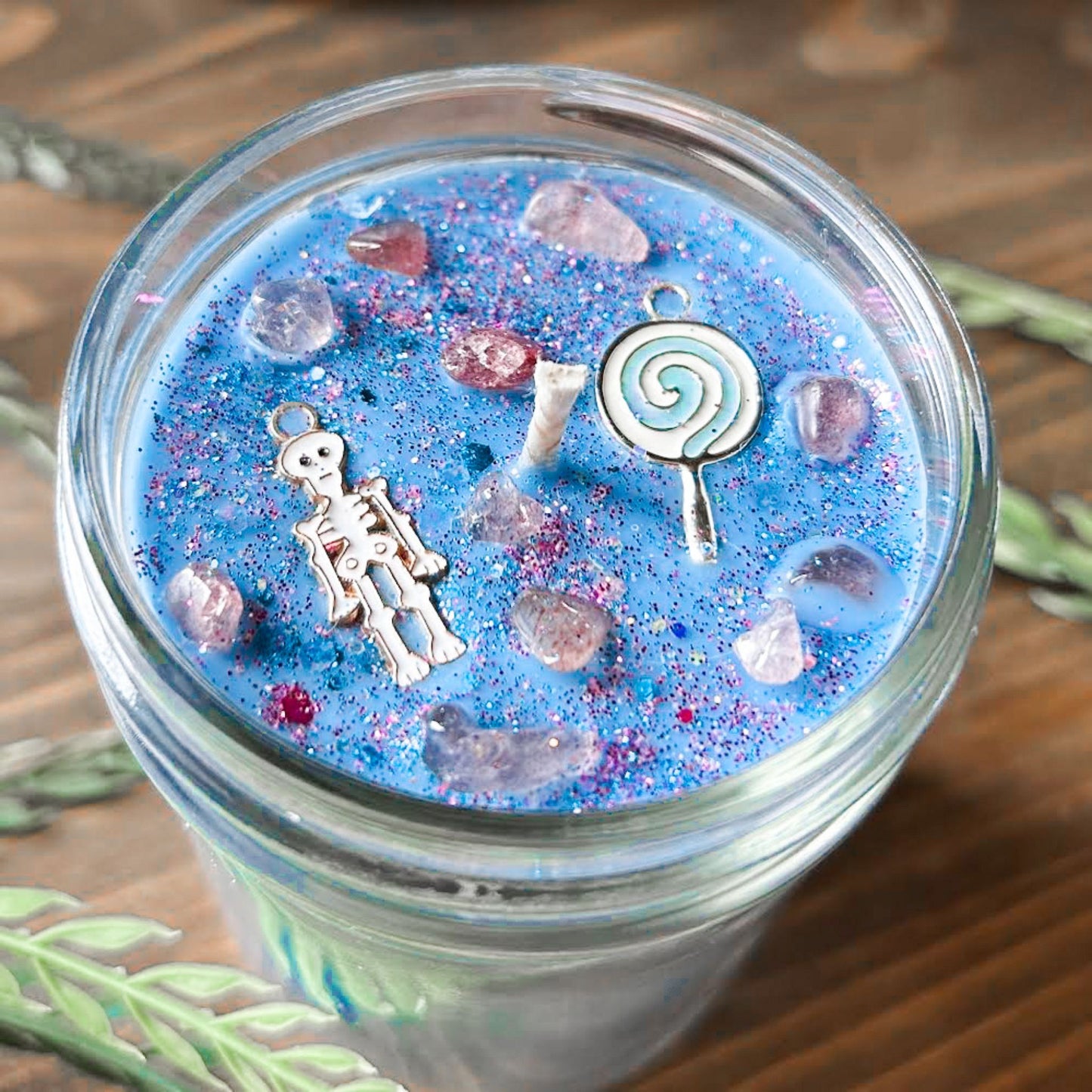 A pastel blue Halloween candle with a creepy cute skeleton, a swirl lollipop, spooky crystals, and gothic pink glitter.