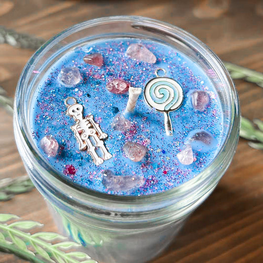A pastel blue Halloween candle with a creepy cute skeleton, a swirl lollipop, spooky crystals, and gothic pink glitter.