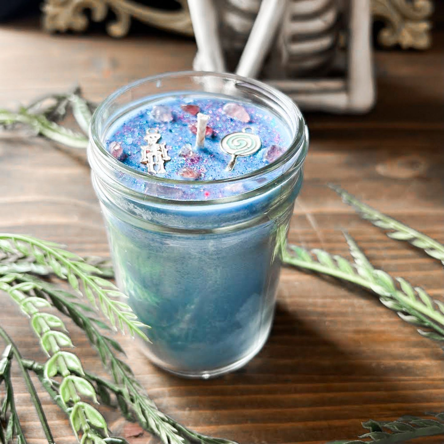 A pastel blue Halloween candle with a creepy cute skeleton, a swirl lollipop, spooky crystals, and gothic pink glitter.