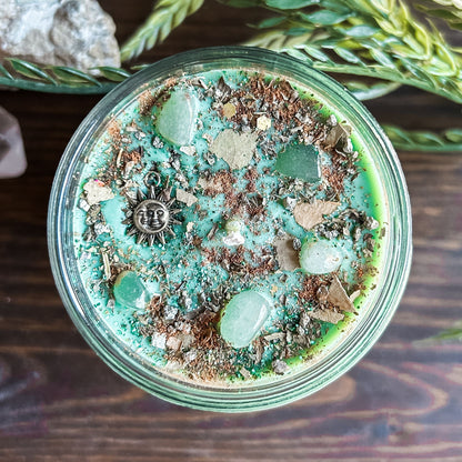 Green intention crystal candle that is decorated with a witchy sun and moon charm, gold magic glitter. The candle also has crystals and herbs for manifesting abundance.
