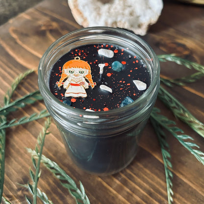 A black Halloween candle that is decorated with an Annabelle horror movie charm, spooky crystals, and paranormal glitter.