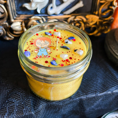 A yellow Halloween candle that is decorated with a horror movie chucky charm, spooky crystals, and creepy glitter.