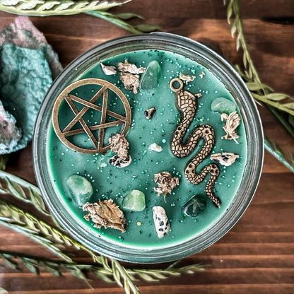 A green witchy candle with a forest aesthetic. The candle has a pentagram and a snake, as well as forest with crystals and spooky earthy herbs.