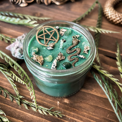 A green witchy candle with a forest aesthetic. The candle has a pentagram and a snake, as well as forest with crystals and spooky earthy herbs.
