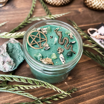 A green witchy candle with a forest aesthetic. The candle has a pentagram and a snake, as well as forest with crystals and spooky earthy herbs.