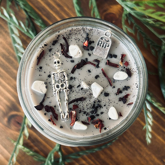A dark Halloween candle that is decorated with a scary skeleton, a spooky rest in peace sign, white crystals, and gothic black glitter.