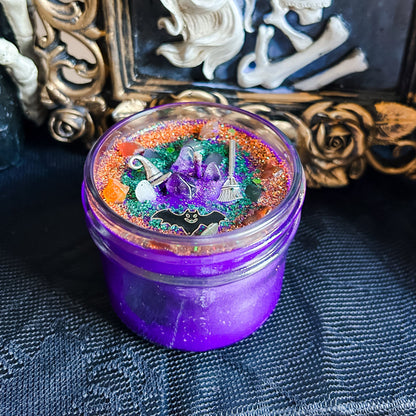 A purple Halloween candle that is decorated with a witch's broom, a witch hat, a spooky bat, witchy crystals, and magic glitter.
