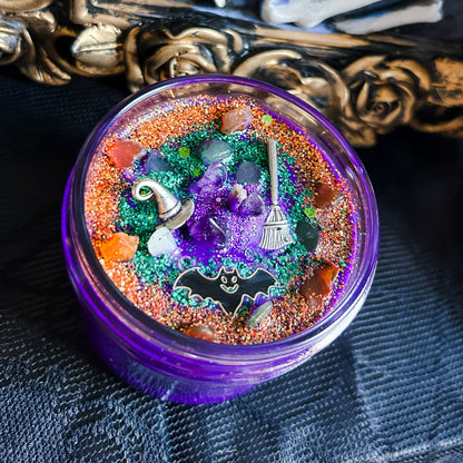 A purple Halloween candle that is decorated with a witch's broom, a witch hat, a spooky bat, witchy crystals, and magic glitter.