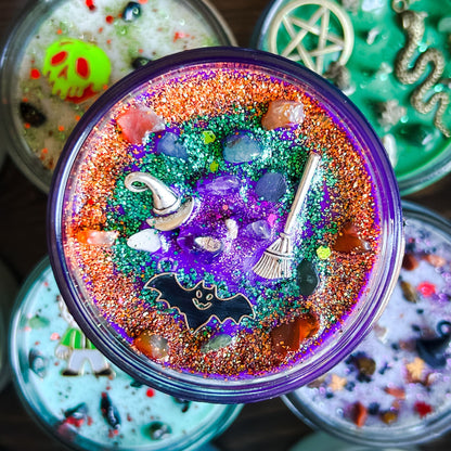A purple Halloween candle that is decorated with a witch's broom, a witch hat, a spooky bat, witchy crystals, and magic glitter.
