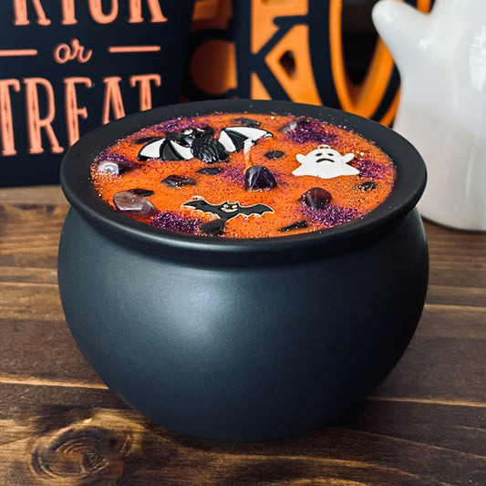 Orange Halloween candle in a black cauldron that is decorated with witchy crystals, spooky bats, and gothic glitter.