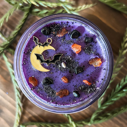 A purple Halloween candle that is decorated with a spooky moon and bats, orange and black crystals, and eerie glitter.