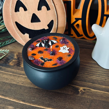 Orange Halloween candle in a black cauldron that is decorated with witchy crystals, spooky bats, and gothic glitter.