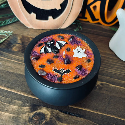 Orange Halloween candle in a black cauldron that is decorated with witchy crystals, spooky bats, and gothic glitter.