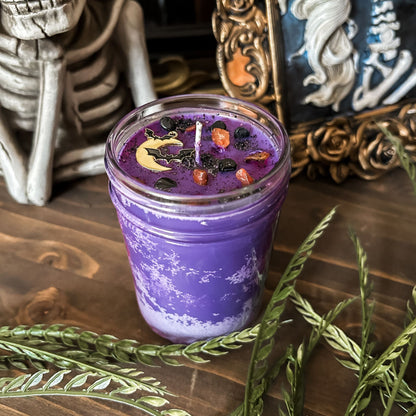 A purple Halloween candle that is decorated with a spooky moon and bats, orange and black crystals, and eerie glitter.