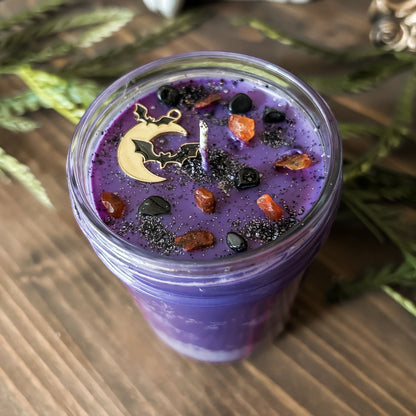 A purple Halloween candle that is decorated with a spooky moon and bats, orange and black crystals, and eerie glitter.