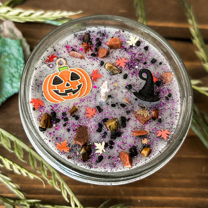 A Halloween candle that is decorated with a jack o lantern, a witch hat, spooky crystals, and autumn glitter.