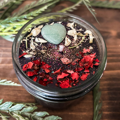 Black intention candle that is decorated with a green aventurine crystal heart. The candle also has jasmine, rose petals, witch hazel, and magic glitter for manifesting.