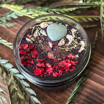 Black intention candle that is decorated with a green aventurine crystal heart. The candle also has jasmine, rose petals, witch hazel, and magic glitter for manifesting.