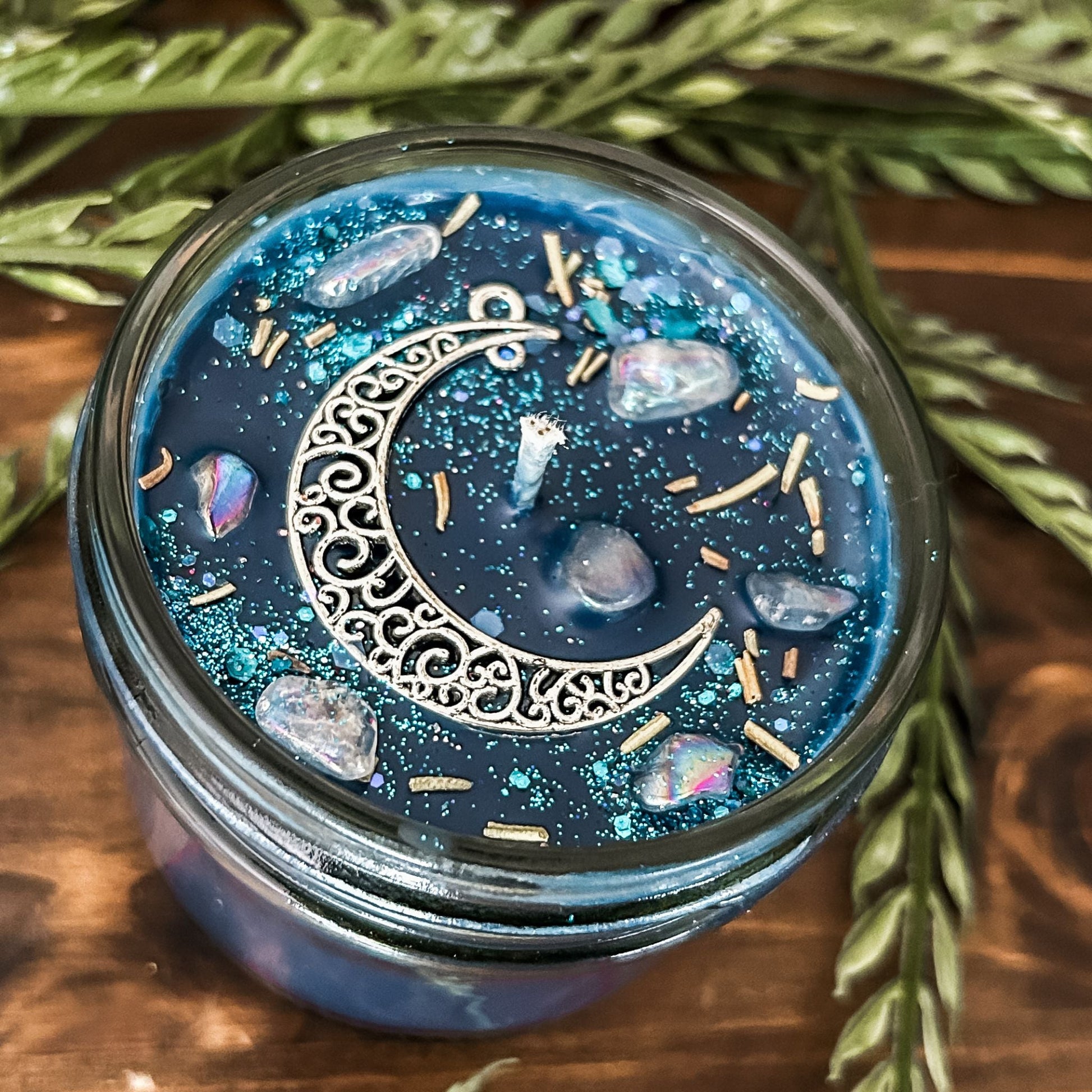 Blue intention candle that is decorated with a silver crescent moon. The candle also has rosemary, aura quartz crystals, and celestial glitter for moon ritual spells and manifesting.