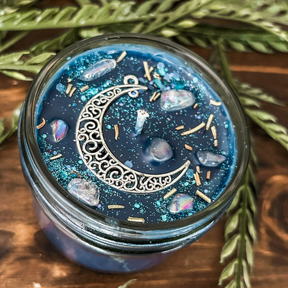 Blue intention candle that is decorated with a silver crescent moon. The candle also has rosemary, aura quartz crystals, and celestial glitter for moon ritual spells and manifesting.