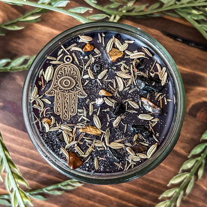 Black crystal intention candle that is decorated with a Hamsa hand, tiger's eye crystal, black tourmaline crystal, thyme, fennel seed, and witchy glitter for protection and grounding.