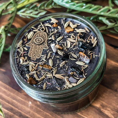 Black crystal intention candle that is decorated with a Hamsa hand, tiger's eye crystal, black tourmaline crystal, thyme, fennel seed, and witchy glitter for protection and grounding.