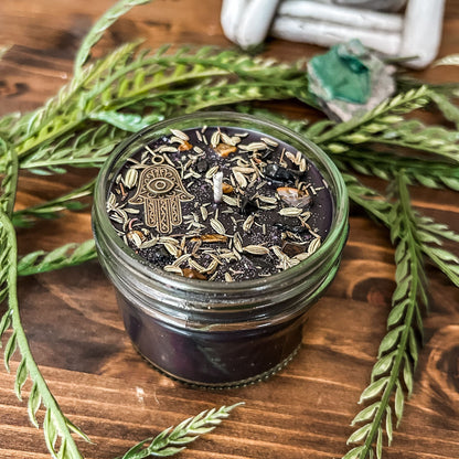 Black crystal intention candle that is decorated with a Hamsa hand, tiger's eye crystal, black tourmaline crystal, thyme, fennel seed, and witchy glitter for protection and grounding.