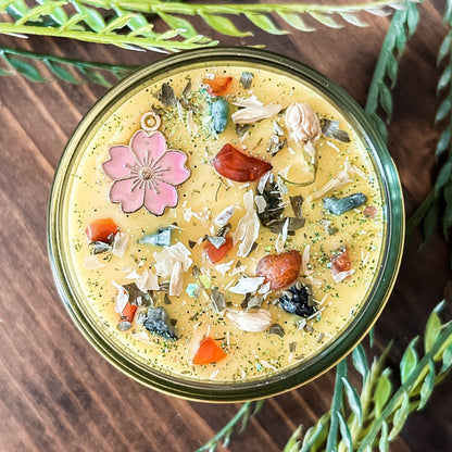 Yellow intention pagan candle that is decorated with a pink flower, crystals, flowers, and magic glitter. The candle represents Beltane and is for setting intentions for renewal, positive energy, purity, and fertility.