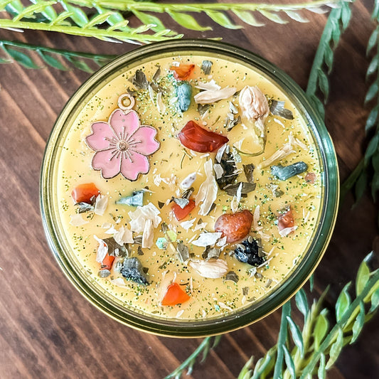 Yellow intention pagan candle that is decorated with a pink flower, crystals, flowers, and magic glitter. The candle represents Beltane and is for setting intentions for renewal, positive energy, purity, and fertility.