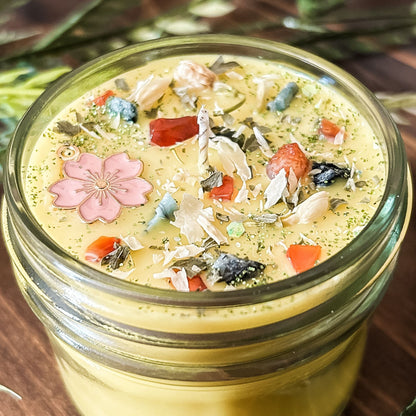 yellow intention pagan candle that is decorated with a pink flower, crystals, flowers, and magic glitter. The candle represents Beltane and is for setting intentions for renewal, positive energy, purity, and fertility.
