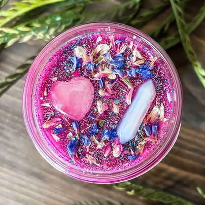 Bright pink witchy intention candle that is decorated with a rose quartz crystal heart, an aura quartz crystal point, flowers, and glitter for manifesting self-love and confidence.