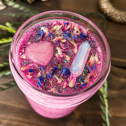 Bright pink witchy intention candle that is decorated with a rose quartz crystal heart, an aura quartz crystal point, flowers, and blue glitter for manifesting self-love and confidence.