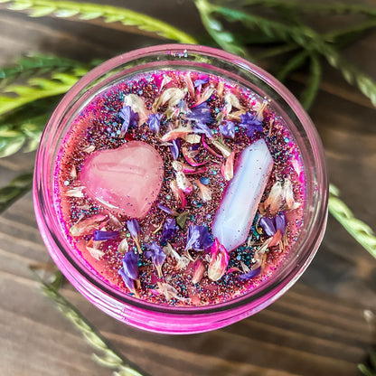 Bright pink witchy intention candle that is decorated with a rose quartz crystal heart, an aura quartz crystal point, flowers, and blue glitter for manifesting self-love and confidence.