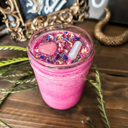Bright pink witchy intention candle that is decorated with a rose quartz crystal heart, an aura quartz crystal point, flowers, and blue glitter for manifesting self-love and confidence.